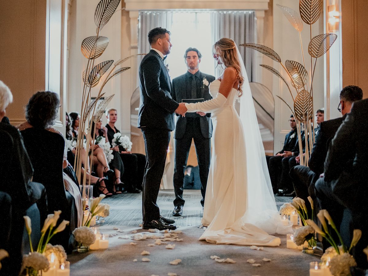 Wedding ceremony inside a hall: a couple holds hands and says vows on a flower-strewn aisle as guests watch.