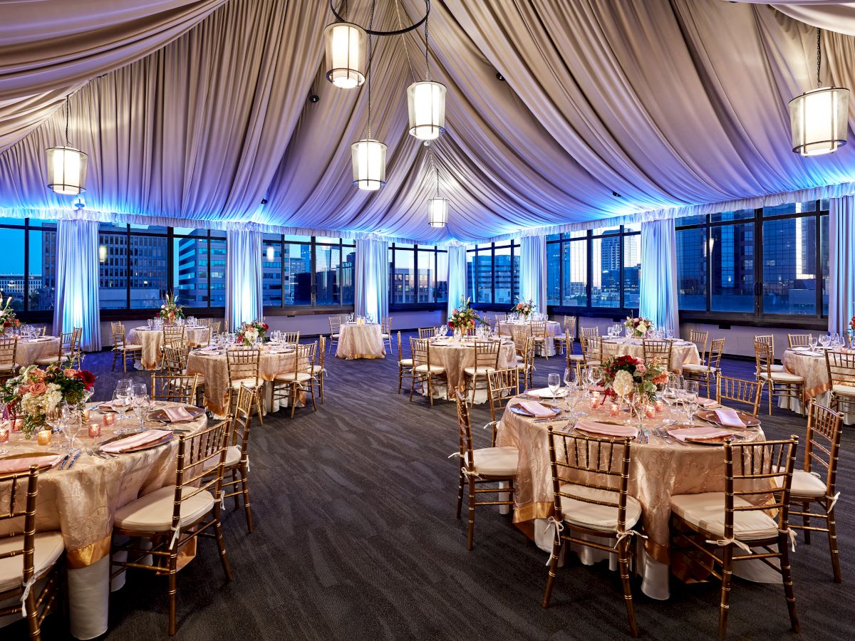 An elegant banquet setup with gold chiavari chairs, floral centerpieces, candle-lit tables, and draped ceiling lamps inside a hall.