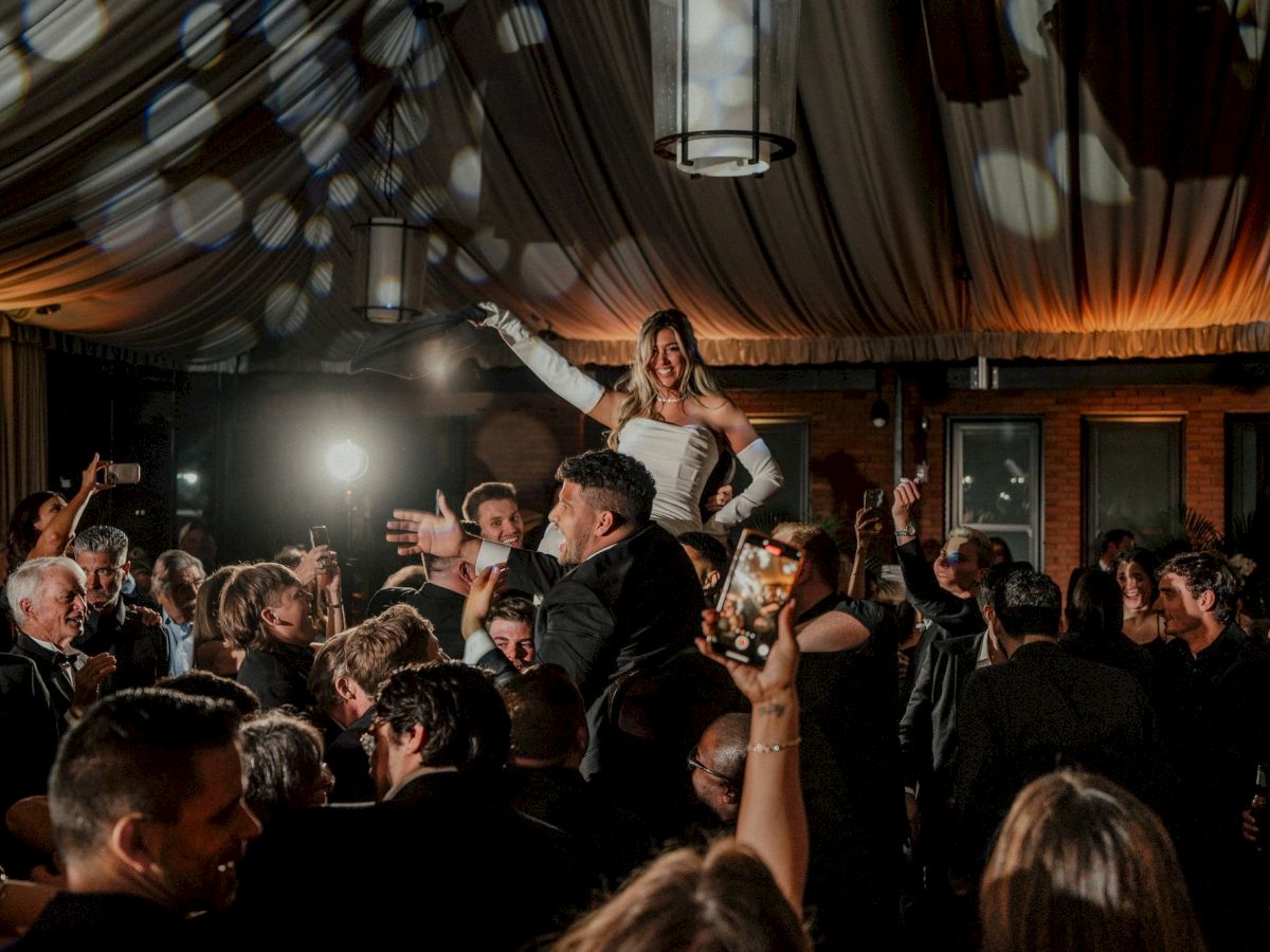 A woman on someone&rsquo;s shoulders celebrates at a crowded wedding reception, smiling under hanging lights and cheers.