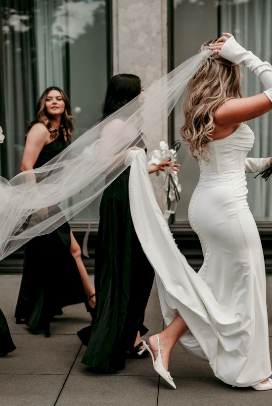 A bride in a white gown and veil twirls outside as bridesmaids in black dresses cheer and carry white orchids.