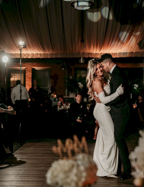 A wedding couple dances on a lit venue dance floor as guests clap in an indoor brick hall, celebrating love.