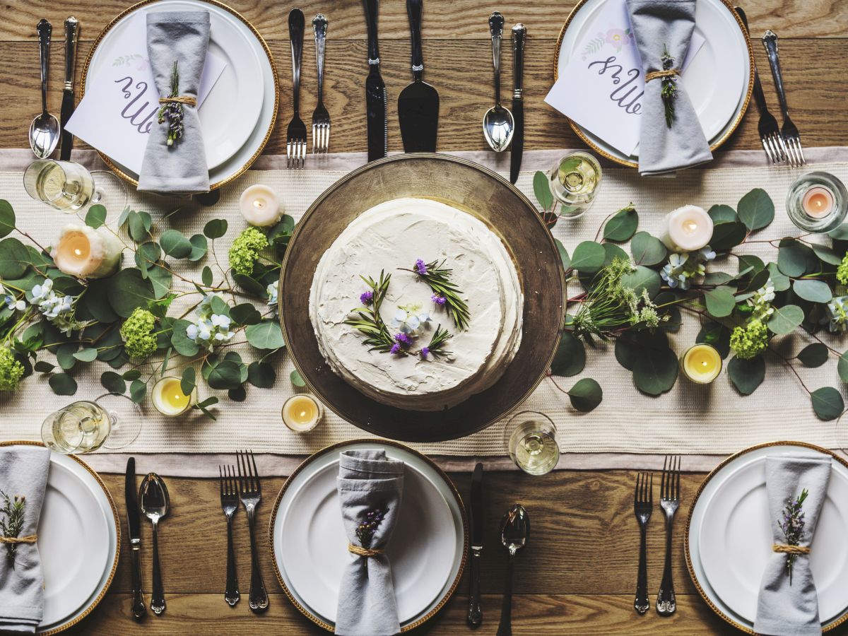 A styled celebration table with plates, cutlery, candles, eucalyptus greenery, and a round frosted cake centerpiece.