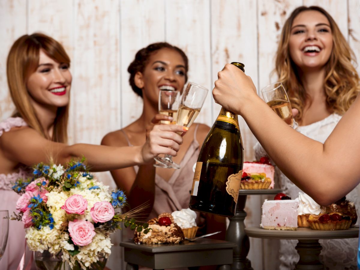 Friends toast with champagne at a celebratory table as flowers and cake sit nearby, smiling and cheering together.