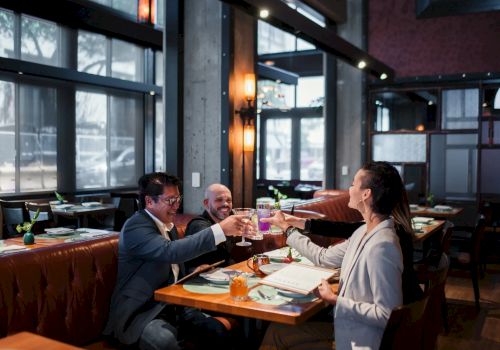 Three people in a modern restaurant toast with glasses, smiling and celebrating together over drinks and dinner.
